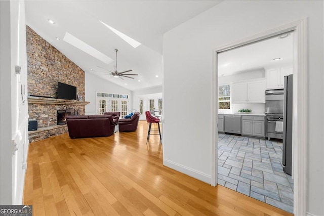 living room featuring ceiling fan, a stone fireplace, a skylight, light wood-style floors, and high vaulted ceiling