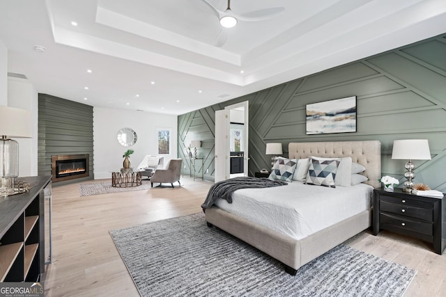 bedroom featuring a large fireplace, light hardwood / wood-style floors, ceiling fan, and a tray ceiling