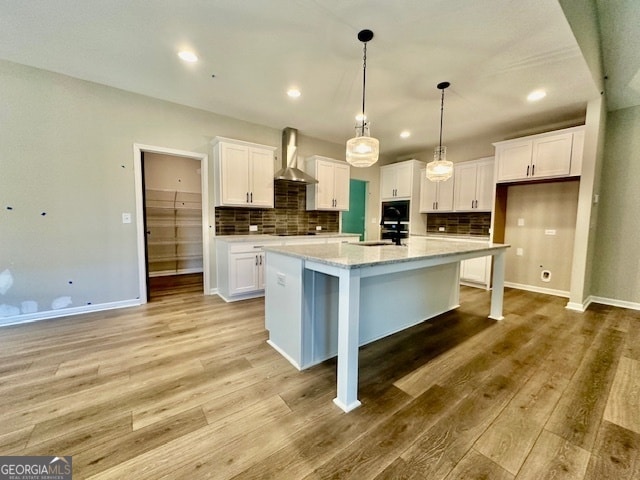 kitchen with wall chimney range hood, backsplash, an island with sink, white cabinetry, and light hardwood / wood-style floors