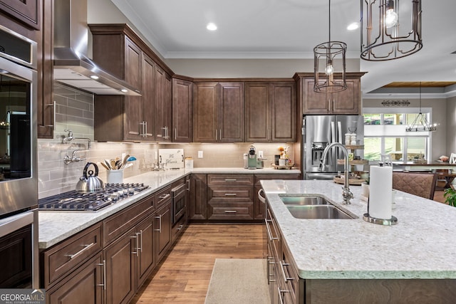 kitchen featuring appliances with stainless steel finishes, sink, wall chimney range hood, pendant lighting, and a center island with sink