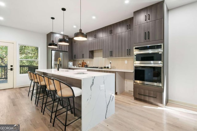 kitchen with an island with sink, stainless steel appliances, decorative light fixtures, light stone counters, and light hardwood / wood-style flooring