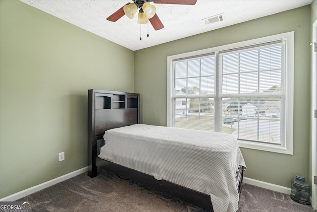 bedroom with a textured ceiling, a ceiling fan, visible vents, baseboards, and dark colored carpet