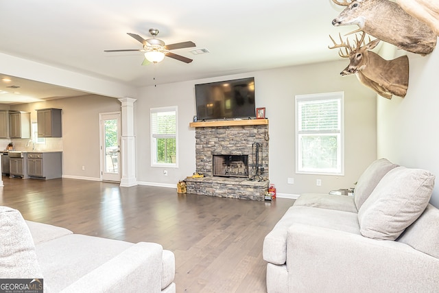 living room with a stone fireplace, ornate columns, dark wood-type flooring, sink, and ceiling fan