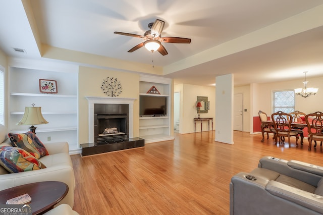 living room with ceiling fan with notable chandelier, built in shelves, hardwood / wood-style flooring, a tile fireplace, and a tray ceiling