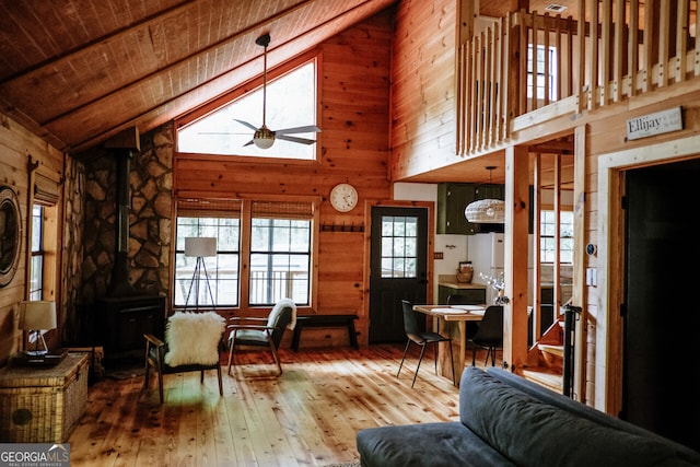 living room featuring wood walls, a wood stove, hardwood / wood-style floors, ceiling fan, and high vaulted ceiling