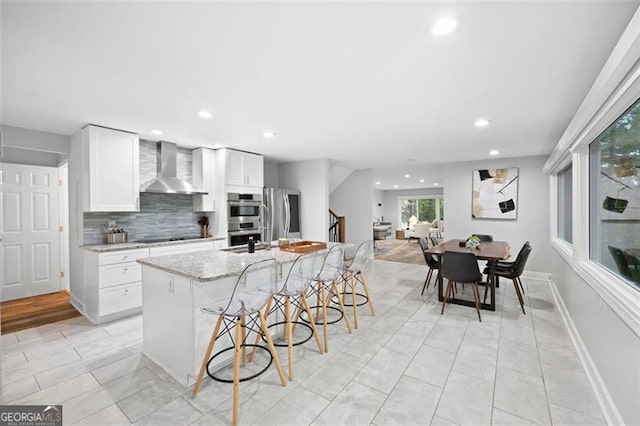 kitchen with wall chimney range hood, appliances with stainless steel finishes, a center island with sink, and white cabinets