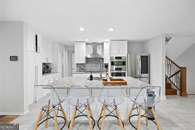 kitchen featuring appliances with stainless steel finishes, a kitchen breakfast bar, wall chimney exhaust hood, light stone counters, and a kitchen island with sink