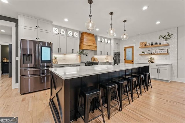 kitchen with stainless steel appliances, a center island with sink, white cabinetry, premium range hood, and light hardwood / wood-style floors