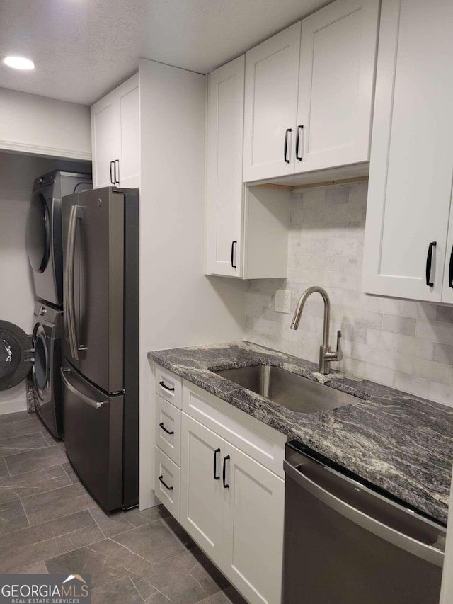 kitchen featuring appliances with stainless steel finishes, backsplash, white cabinetry, dark stone countertops, and stacked washer and clothes dryer