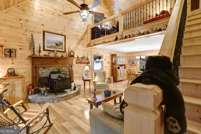 living room featuring a wood stove, high vaulted ceiling, ceiling fan with notable chandelier, wooden walls, and light hardwood / wood-style flooring