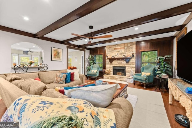 living area featuring arched walkways, a stone fireplace, wood finished floors, a ceiling fan, and beamed ceiling