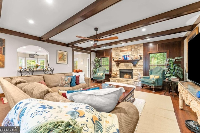 living room featuring arched walkways, a stone fireplace, wood finished floors, a ceiling fan, and beamed ceiling