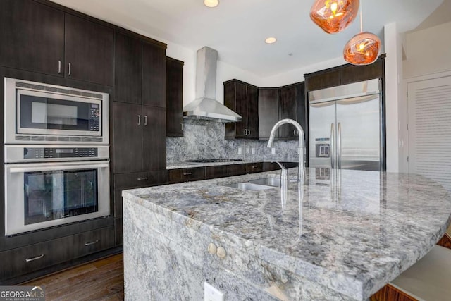 kitchen featuring wall chimney range hood, built in appliances, light stone counters, dark hardwood / wood-style floors, and a kitchen island with sink