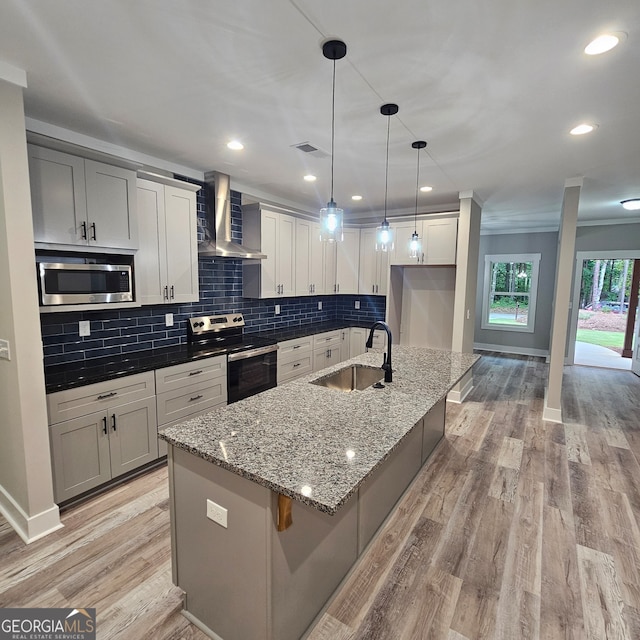 kitchen with wall chimney range hood, sink, light wood-type flooring, appliances with stainless steel finishes, and an island with sink