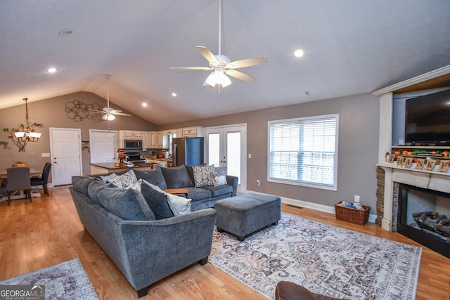 living room with ceiling fan with notable chandelier, a stone fireplace, light wood-type flooring, and vaulted ceiling