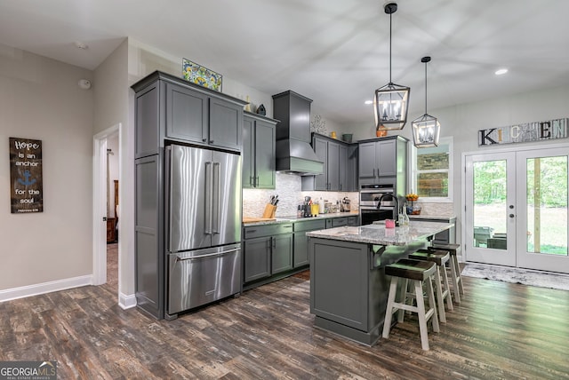 kitchen featuring premium range hood, hanging light fixtures, stainless steel appliances, light stone countertops, and an island with sink