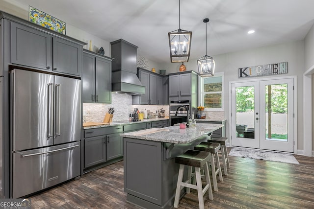 kitchen with appliances with stainless steel finishes, hanging light fixtures, light stone countertops, custom range hood, and a center island with sink