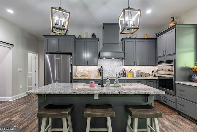 kitchen with appliances with stainless steel finishes, dark hardwood / wood-style floors, light stone countertops, an island with sink, and custom exhaust hood