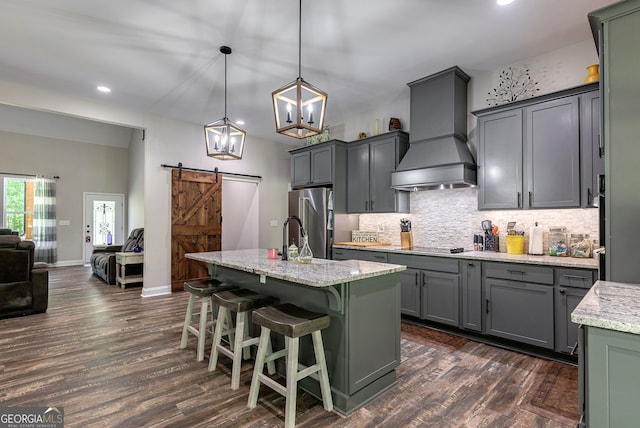 kitchen with hanging light fixtures, high end fridge, a barn door, custom range hood, and a center island with sink