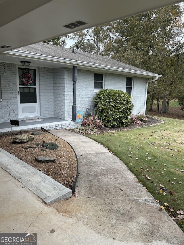 doorway to property featuring brick siding, covered porch, a lawn, and roof with shingles