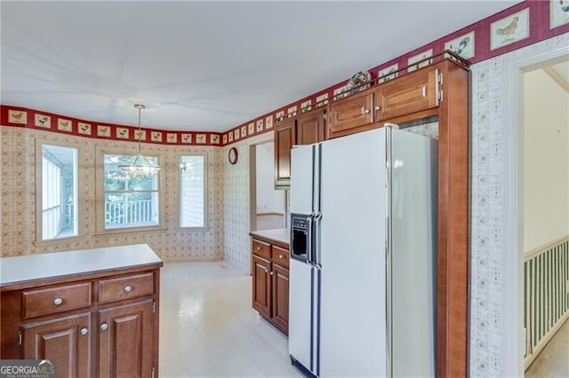 kitchen featuring white refrigerator with ice dispenser, wallpapered walls, light countertops, brown cabinets, and hanging light fixtures