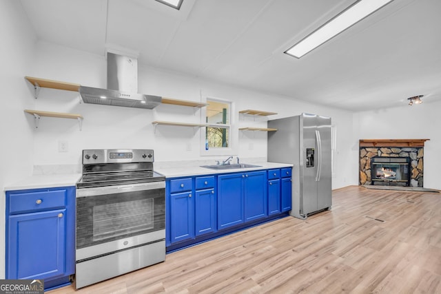 kitchen with blue cabinetry, light hardwood / wood-style flooring, a stone fireplace, wall chimney exhaust hood, and stainless steel appliances