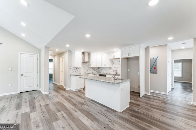 kitchen with light stone countertops, a center island with sink, white cabinets, and wall chimney range hood