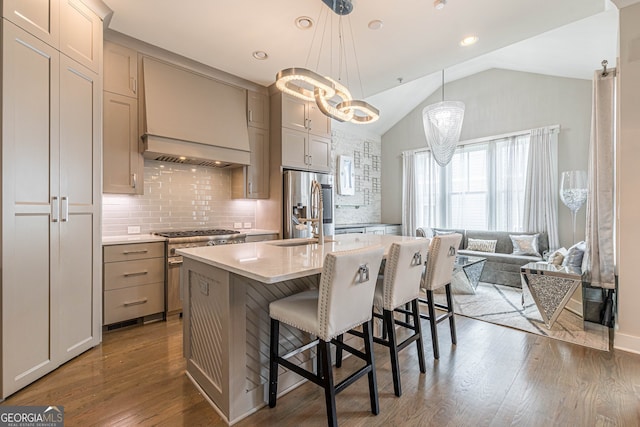 kitchen with appliances with stainless steel finishes, an island with sink, hanging light fixtures, lofted ceiling, and premium range hood