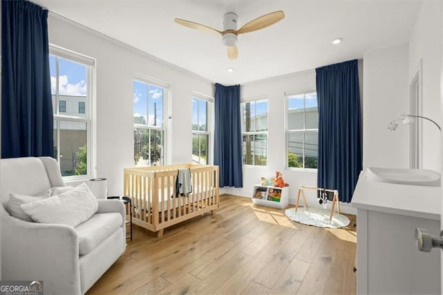 bedroom featuring a crib, ceiling fan, multiple windows, and light wood-type flooring