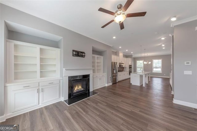 unfurnished living room with built in shelves, ceiling fan with notable chandelier, dark wood-type flooring, and sink