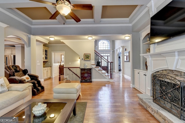 living room featuring ceiling fan, a stone fireplace, crown molding, decorative columns, and light wood-type flooring