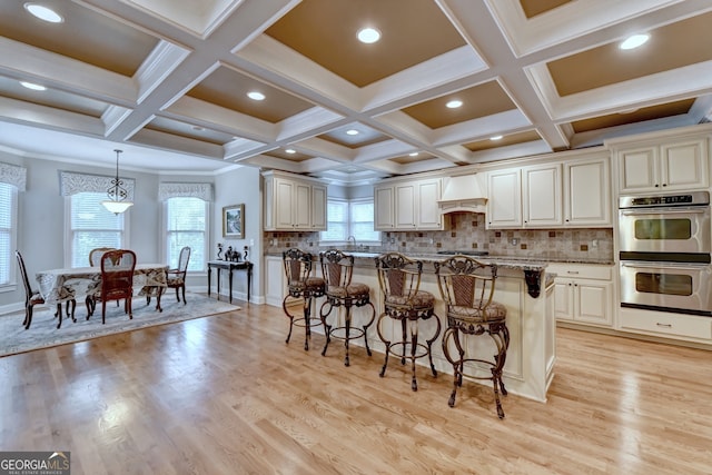 kitchen featuring light stone counters, decorative light fixtures, light wood-type flooring, double oven, and premium range hood