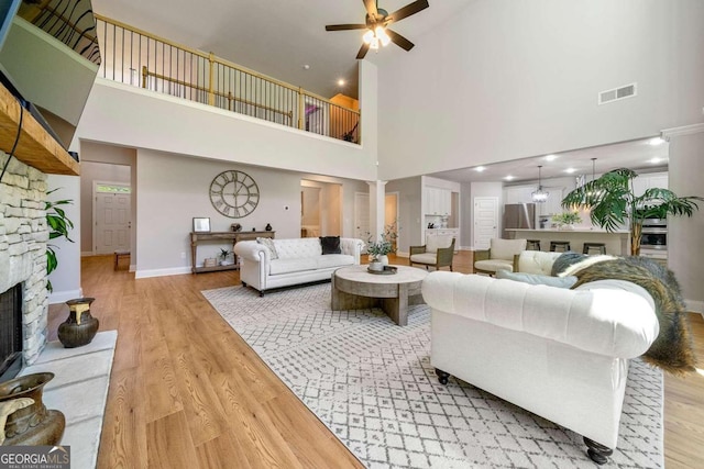 living room with a high ceiling, ceiling fan, wood-type flooring, a stone fireplace, and ornate columns