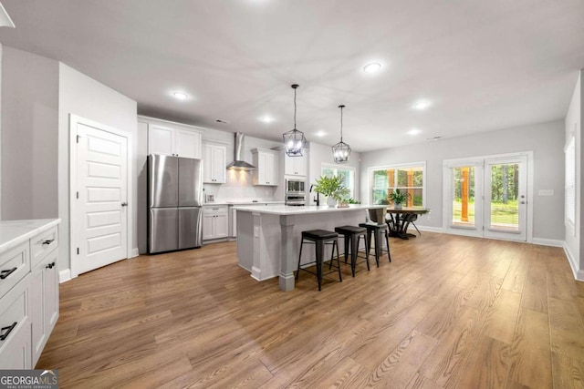 kitchen with stainless steel appliances, hanging light fixtures, an island with sink, light hardwood / wood-style floors, and wall chimney exhaust hood