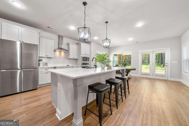 kitchen featuring stainless steel appliances, a kitchen island with sink, wall chimney range hood, white cabinetry, and light hardwood / wood-style flooring