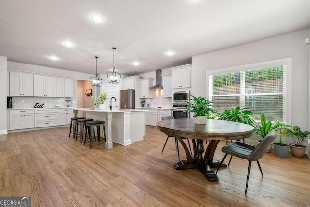 kitchen with appliances with stainless steel finishes, hanging light fixtures, an island with sink, white cabinets, and wall chimney exhaust hood