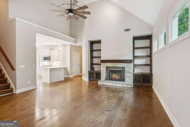 unfurnished living room with ceiling fan, sink, hardwood / wood-style flooring, high vaulted ceiling, and a stone fireplace