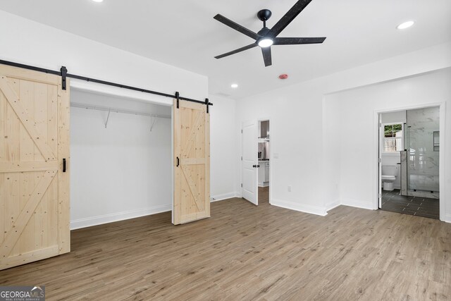 unfurnished bedroom featuring hardwood / wood-style floors, ensuite bathroom, a barn door, and ceiling fan