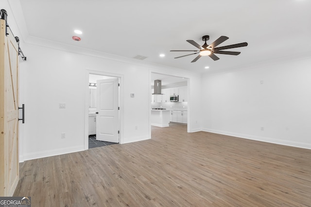 unfurnished living room featuring hardwood / wood-style flooring, a barn door, ceiling fan, and crown molding