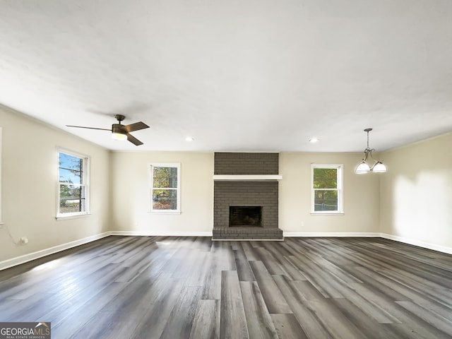 unfurnished living room with a brick fireplace, ceiling fan with notable chandelier, dark hardwood / wood-style flooring, and a healthy amount of sunlight