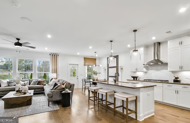 kitchen featuring a center island with sink, wall chimney exhaust hood, white cabinetry, light hardwood / wood-style flooring, and decorative light fixtures