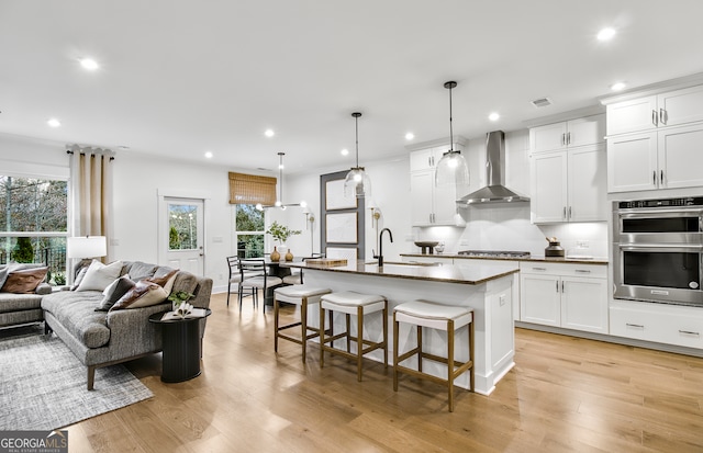 kitchen featuring a center island with sink, white cabinetry, appliances with stainless steel finishes, wall chimney exhaust hood, and hanging light fixtures