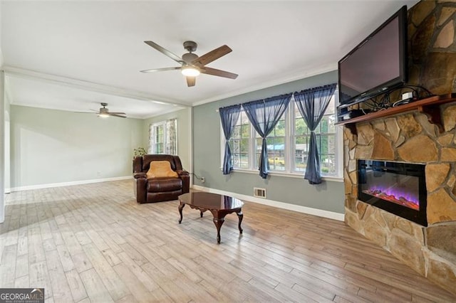 living area featuring a stone fireplace, light wood-type flooring, ceiling fan, and crown molding