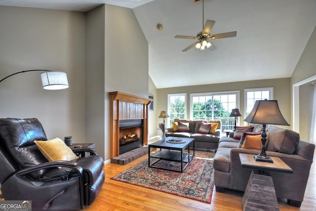 living room with ceiling fan, high vaulted ceiling, wood finished floors, baseboards, and a brick fireplace