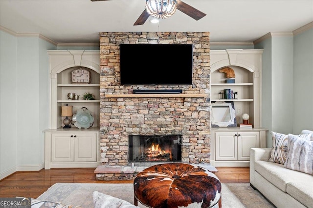 living room with built in shelves, a fireplace, ceiling fan, crown molding, and light wood-type flooring