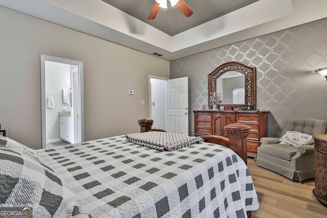 bedroom featuring light hardwood / wood-style floors, ceiling fan, ensuite bath, and a raised ceiling