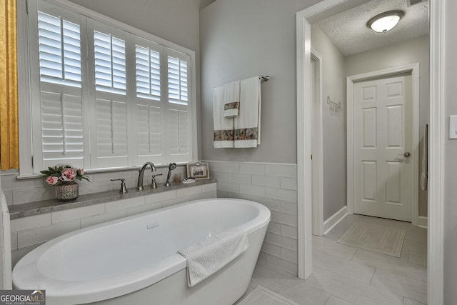 bathroom featuring tile patterned floors, a wainscoted wall, a textured ceiling, tile walls, and a soaking tub