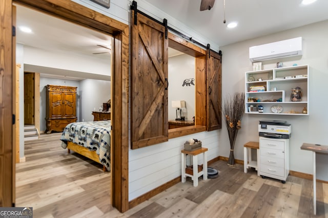 bedroom featuring a wall mounted AC, wood walls, a barn door, ceiling fan, and light wood-type flooring