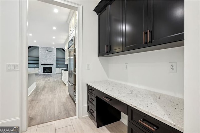 kitchen with a stone fireplace, light hardwood / wood-style floors, built in fridge, light stone counters, and built in desk