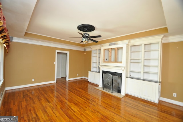 unfurnished living room featuring hardwood / wood-style flooring, built in shelves, a tray ceiling, crown molding, and ceiling fan